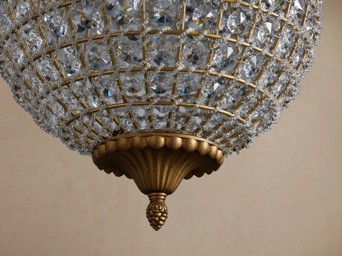 Poppy Chandelier, a spherical fixture covered in clear multifaceted crystals, hanging from an antiqued brass chain against a beige wall.
