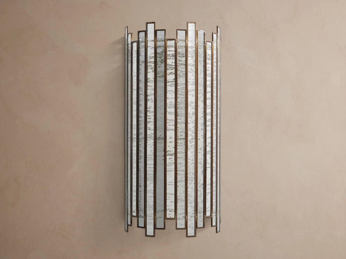 Laila Sconce: vertical antique copper frame with rectangular antiqued mirrored-glass panels, glowing softly against a beige wall.