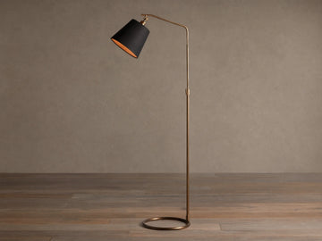 Kellen Antiqued Brass Floor Lamp, slender frame, black fabric shade with warm interior, round base on wooden floor.