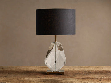 Lola Teardrop Table Lamp with black drum shade, clear faceted crystal base, and brass accents, on a wooden table.
