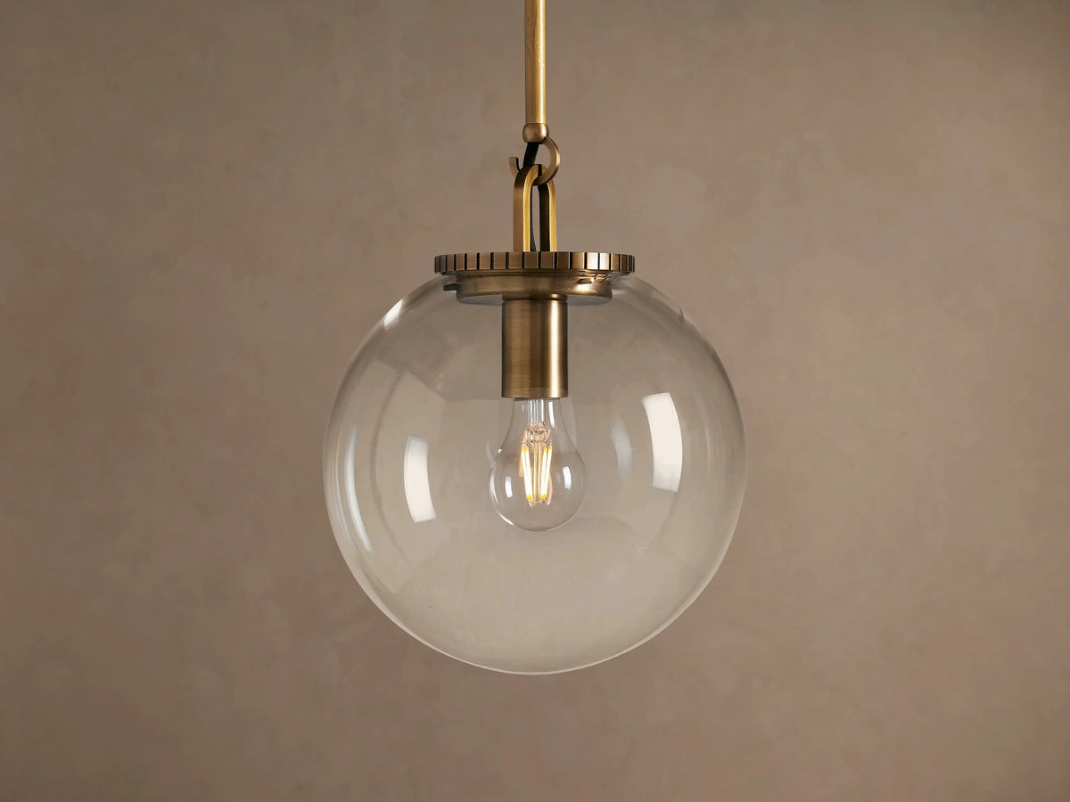 Wescott Globe Pendant: Brass-finished rod and hardware holding a clear glass globe with a filament bulb inside, against a plain wall.