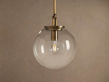 Wescott Globe Pendant: Brass-finished rod and hardware holding a clear glass globe with a filament bulb inside, against a plain wall.