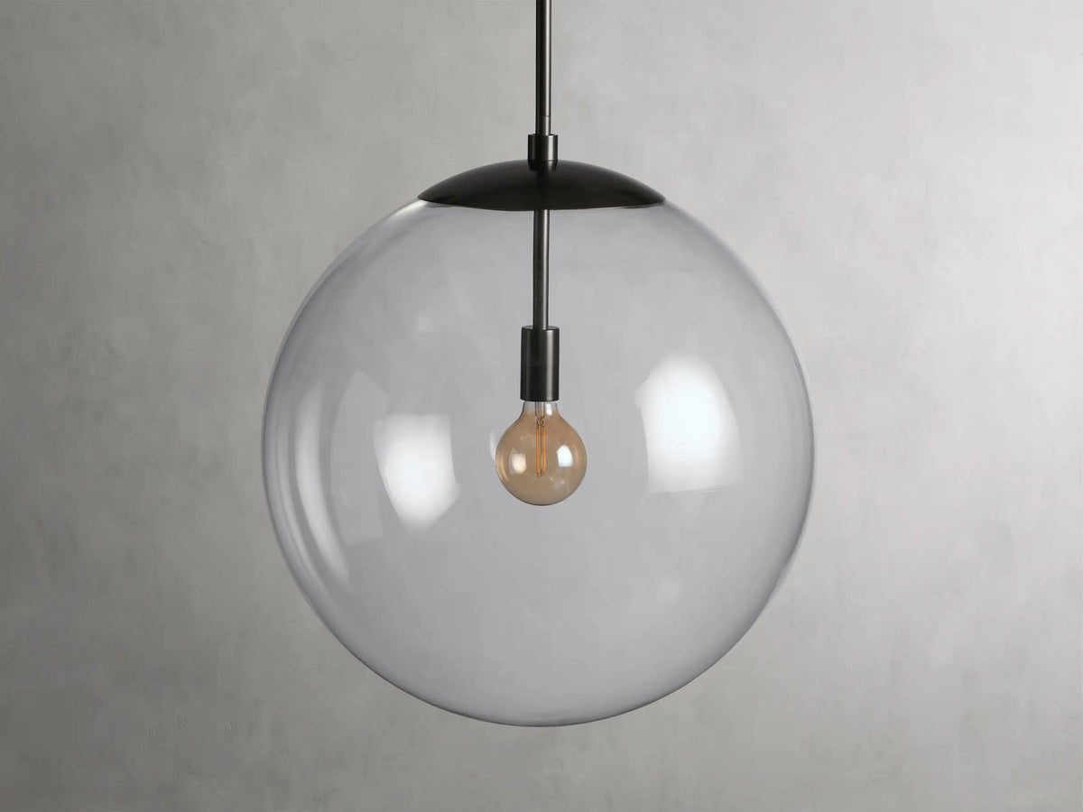 Clear glass globe pendant light with brass rod, exposed bulb, and black fabric-wrapped cord, against gray background.