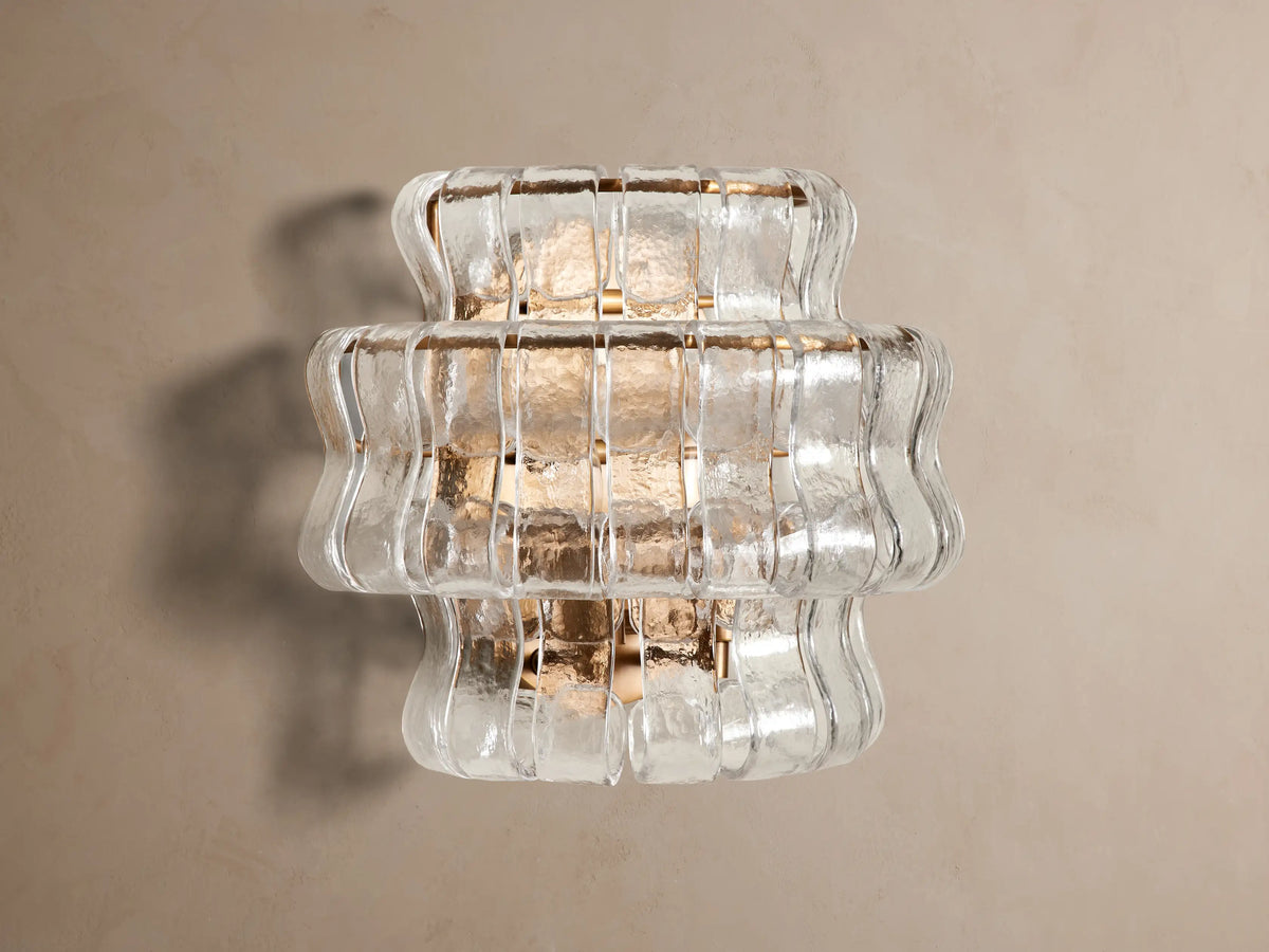 Ghiaccio Sconce: a layered wall light featuring clear, wavy textured glass and a warm, gold-toned frame, illuminated on a wall.