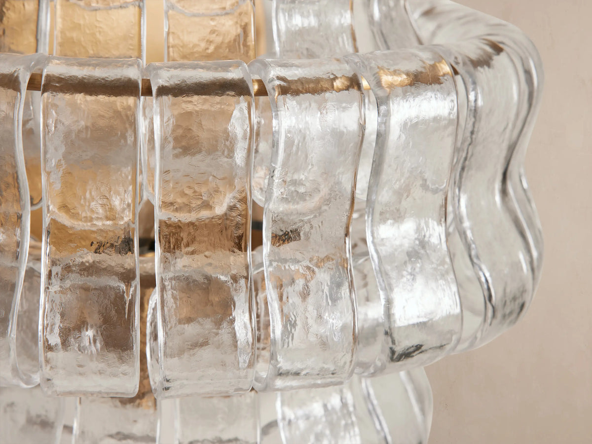 Ghiaccio Sconce: Glowing, textured wavy clear glass and gold-toned steel sconce, vertically oriented on a light beige wall.