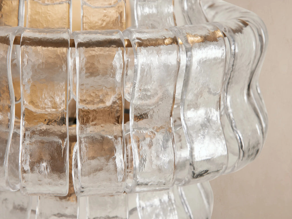 Ghiaccio Sconce: Glowing, textured wavy clear glass and gold-toned steel sconce, vertically oriented on a light beige wall.