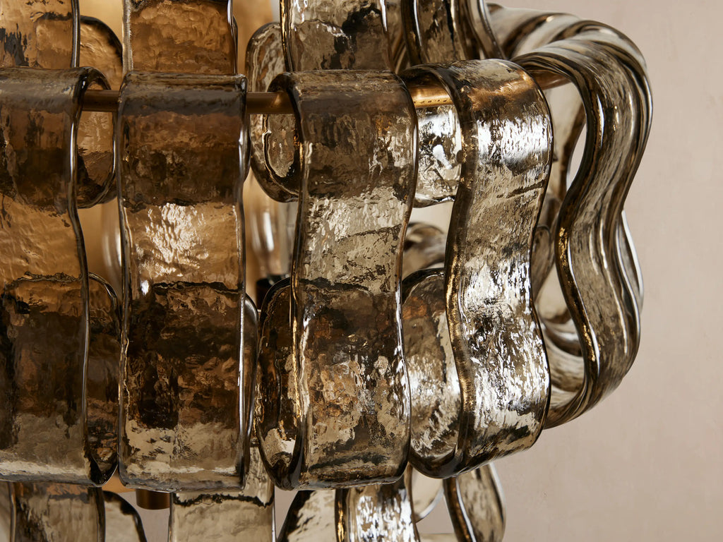 Ghiaccio Sconce, a wall-mounted light with wavy, textured smoky amber glass panels over a brass-toned frame on a neutral wall.
