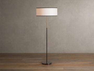 Daphne Floor Lamp with a slim bronze-finished stand, round white linen shade with black trim, wooden floor background.