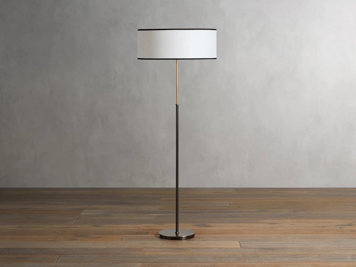 Daphne Floor Lamp with a slim bronze-finished stand, round white linen shade with black trim, wooden floor background.