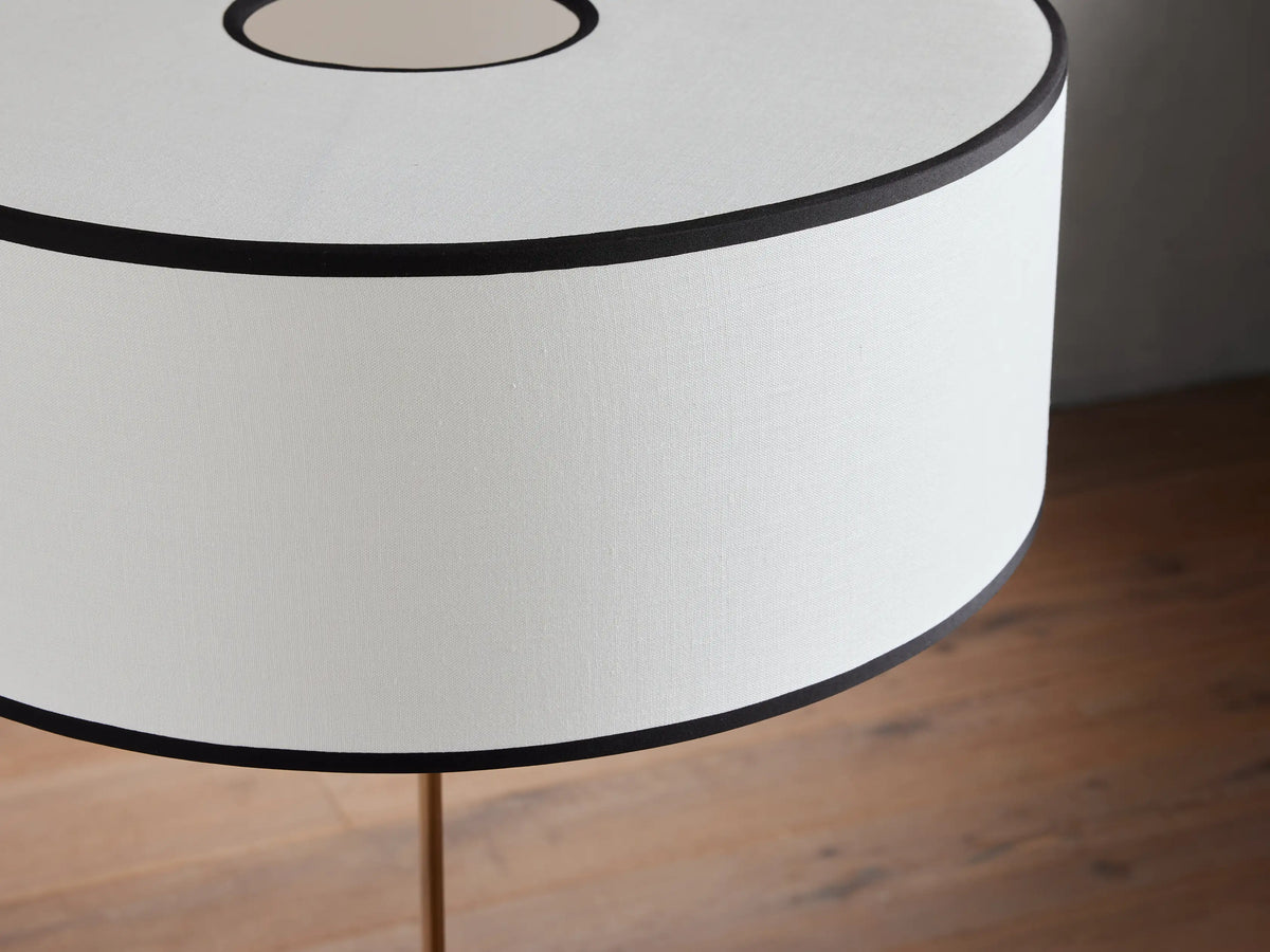 Daphne Floor Lamp with a slender black base and a large white linen shade trimmed in black, standing on a wooden floor.