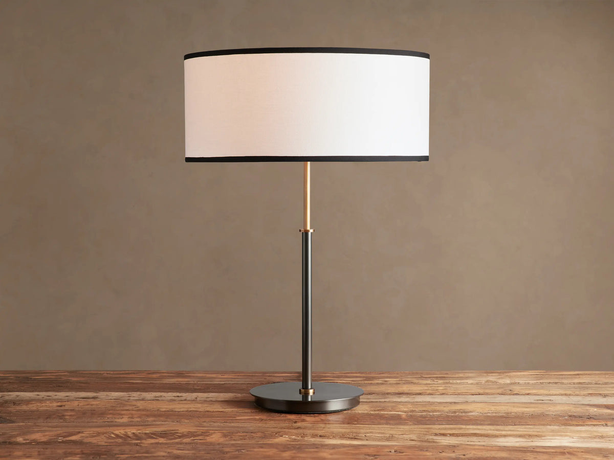 Daphne Table Lamp with white linen shade, black trim, dark bronze finish, and brass accent, on a wooden table.