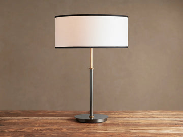 Daphne Table Lamp with white linen shade, black trim, dark bronze finish, and brass accent, on a wooden table.