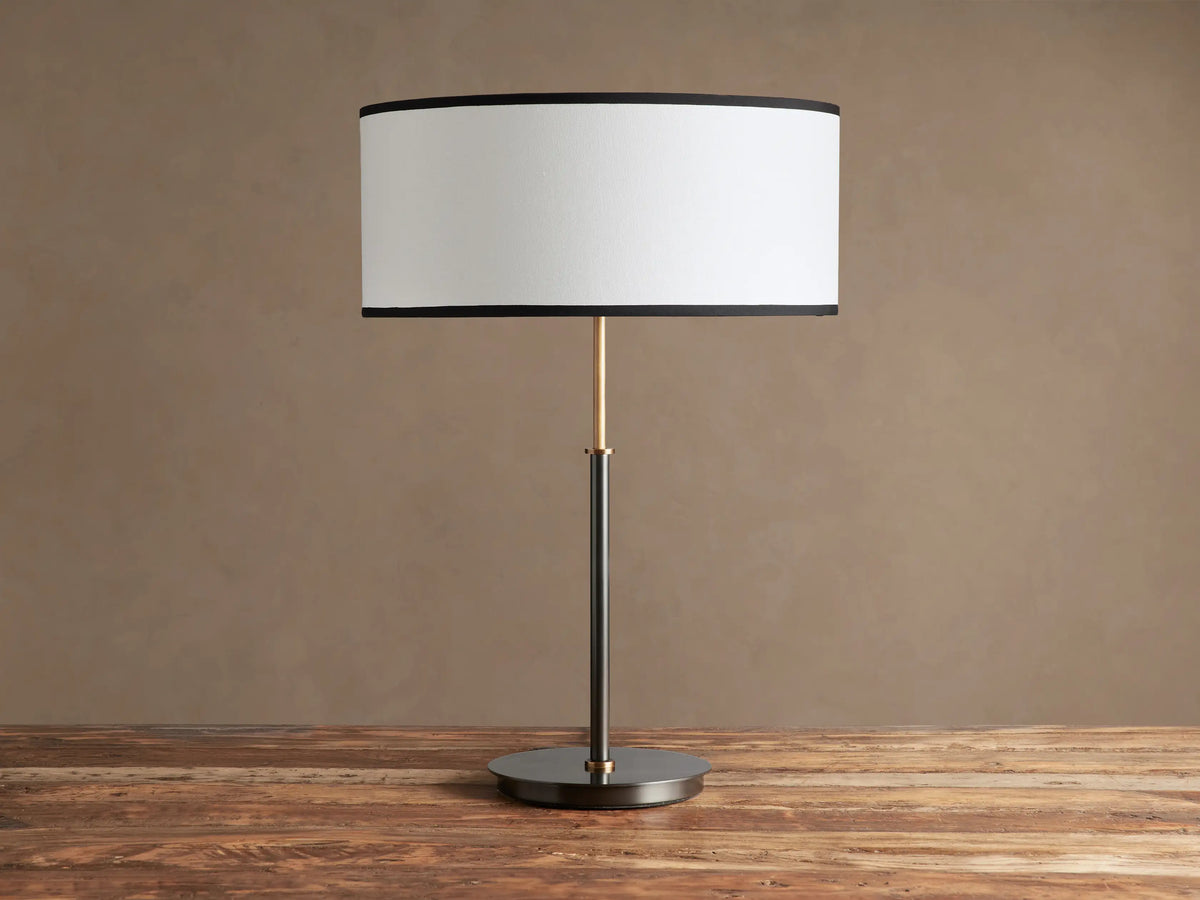Daphne Table Lamp with white linen shade, black trim, dark bronze finish, and brass accent, on a wooden table.