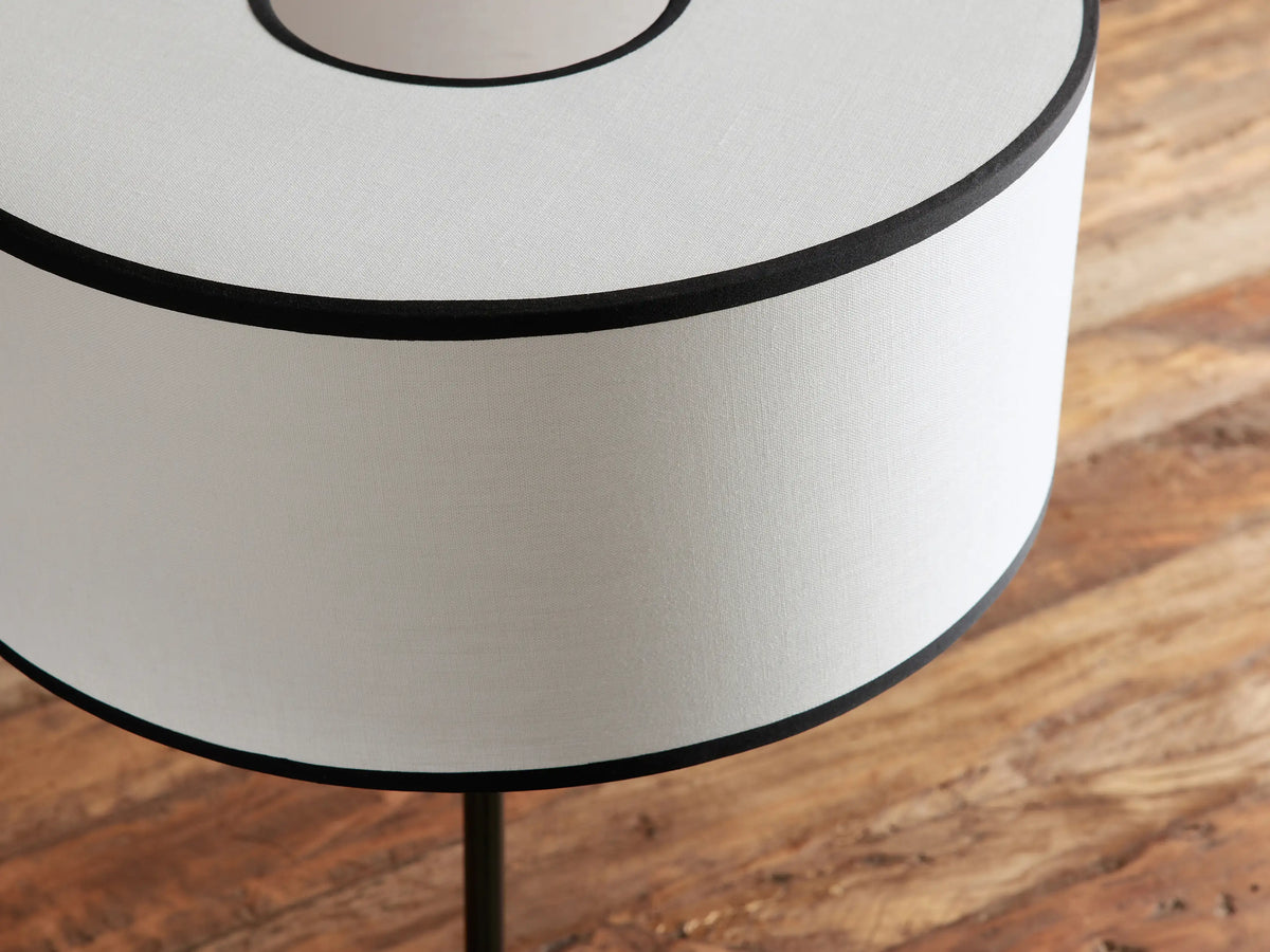 Daphne Table Lamp with a bronze base, gold stem accent, and white linen shade with black trim on a wooden table.
