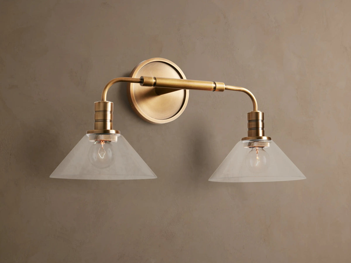 Theron Double Sconce with a brass finish, featuring two milky glass cone shades, horizontally mounted on a taupe wall.