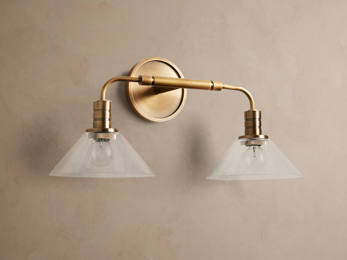 Theron Double Sconce with a brass finish, featuring two milky glass cone shades, horizontally mounted on a taupe wall.