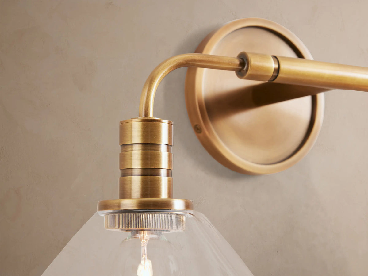 Horizontal Theron Double Sconce in brass with two clear glass cone shades and visible bulbs, mounted on a tan wall.