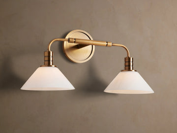 Theron Double Sconce: A brass-finished wall light with two horizontal arms holding conical milk glass shades on a grey wall.
