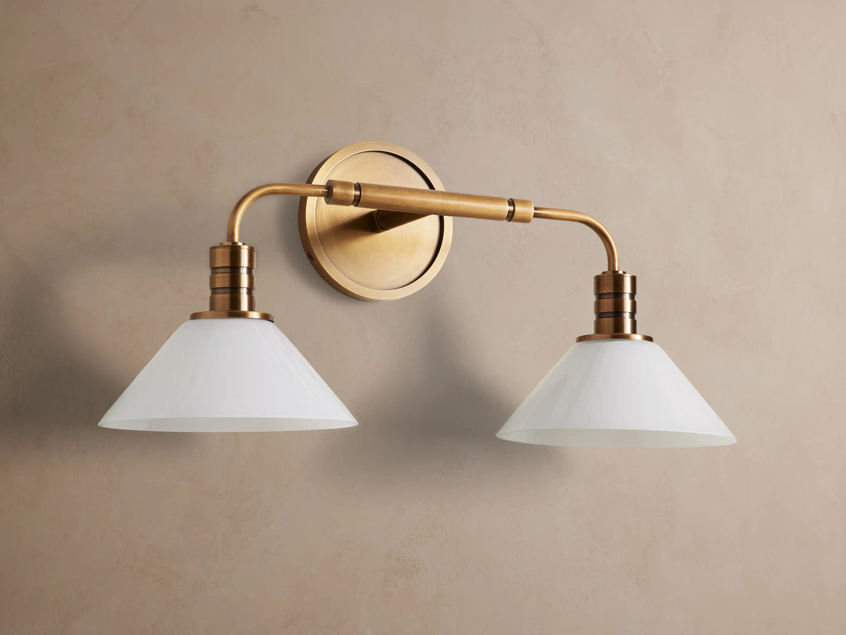 Theron Double Sconce: A brass-finished wall light with two horizontal arms holding conical milk glass shades on a grey wall.