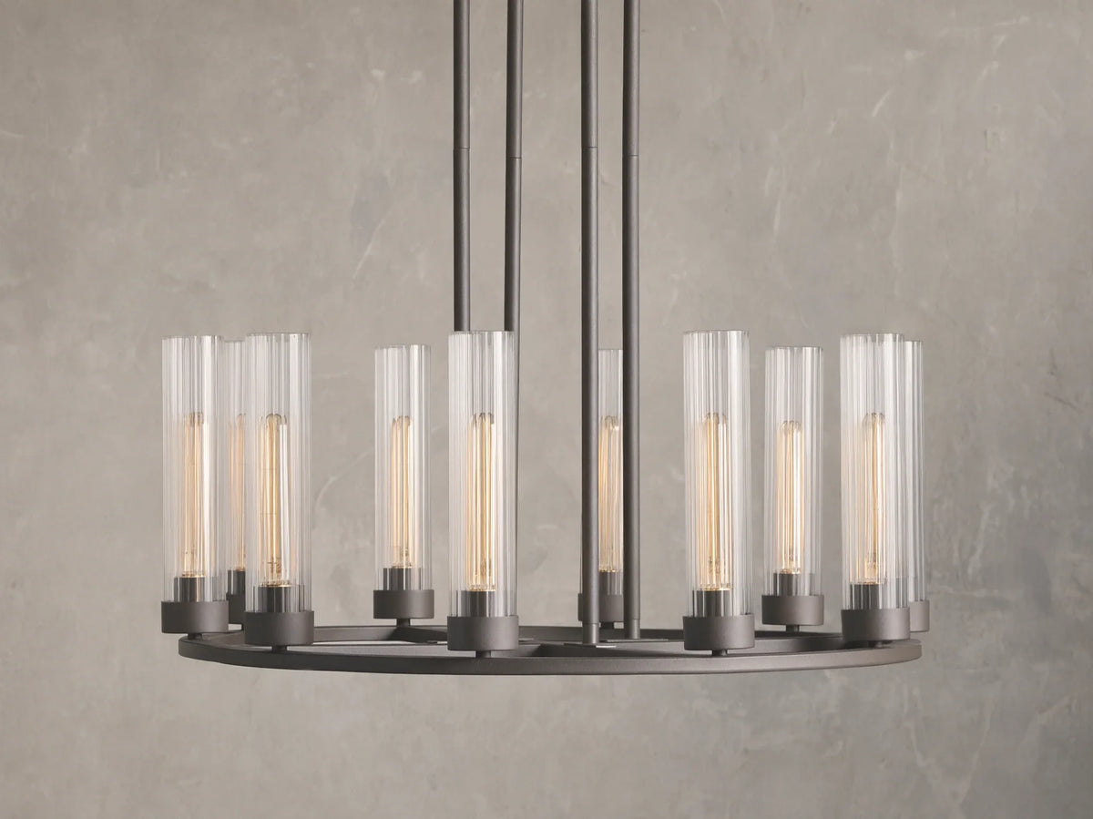 Andreas 10-Light Round Chandelier with a bronzed steel frame, featuring ten vertical, tubular clear glass shades. Mounted against a textured grey wall.