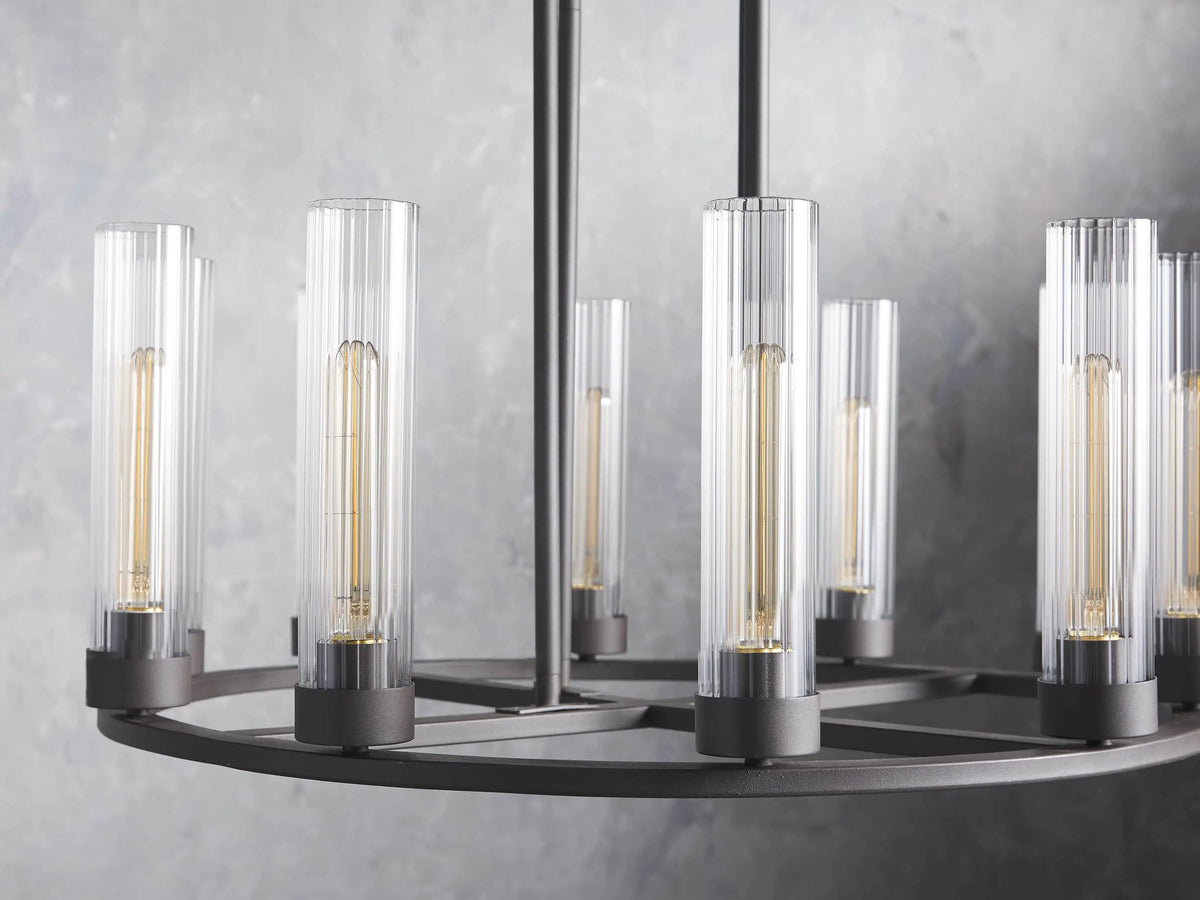 Andreas 10-Light Round Chandelier with a bronzed steel frame, featuring ten vertical, tubular clear glass shades. Mounted against a textured grey wall.