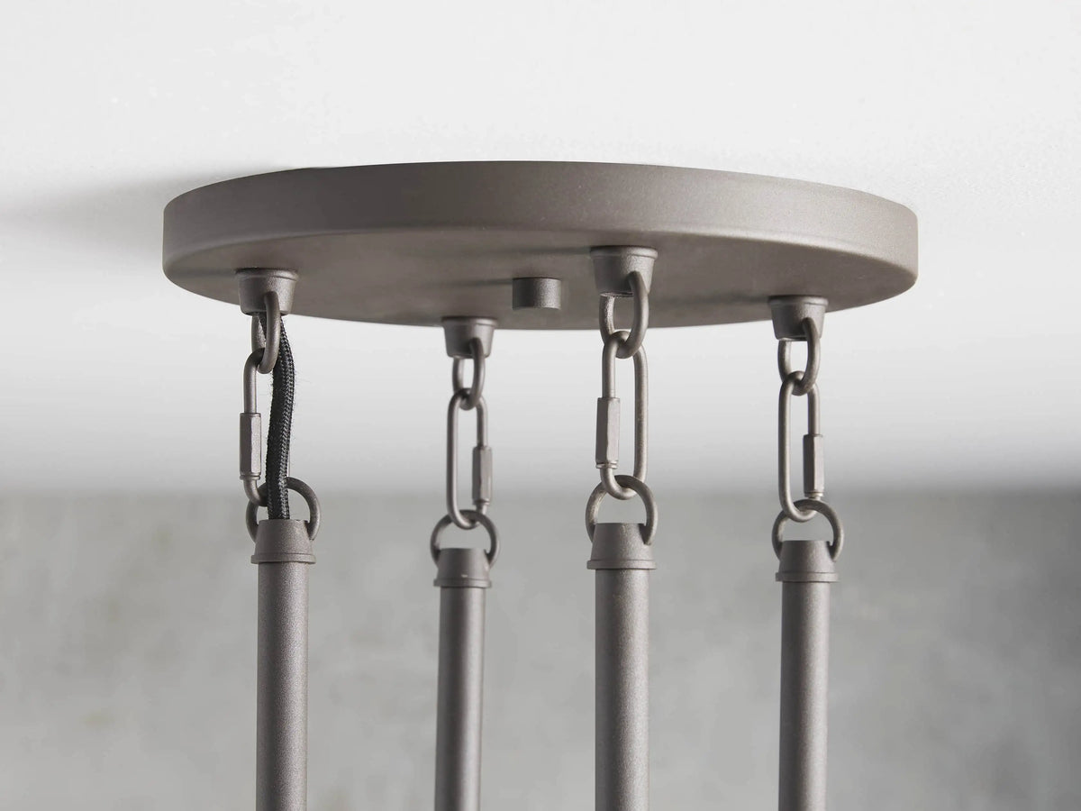 Round chandelier with 10 clear tubular glass shades, black steel frame, vertical rods; shown against a grey textured wall.