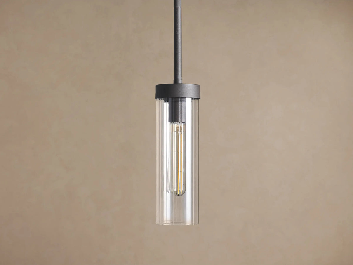 Andreas Pendant: Dark metal rod and cylindrical fixture holding a clear glass shade with a visible vintage filament bulb against a brown wall.