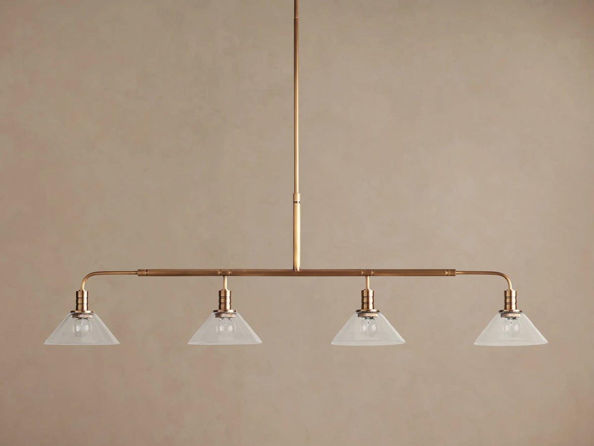 Theron Linear Chandelier: horizontal brass bar, 4 lit bulbs inside milk glass cone shades, hanging against brown wall.