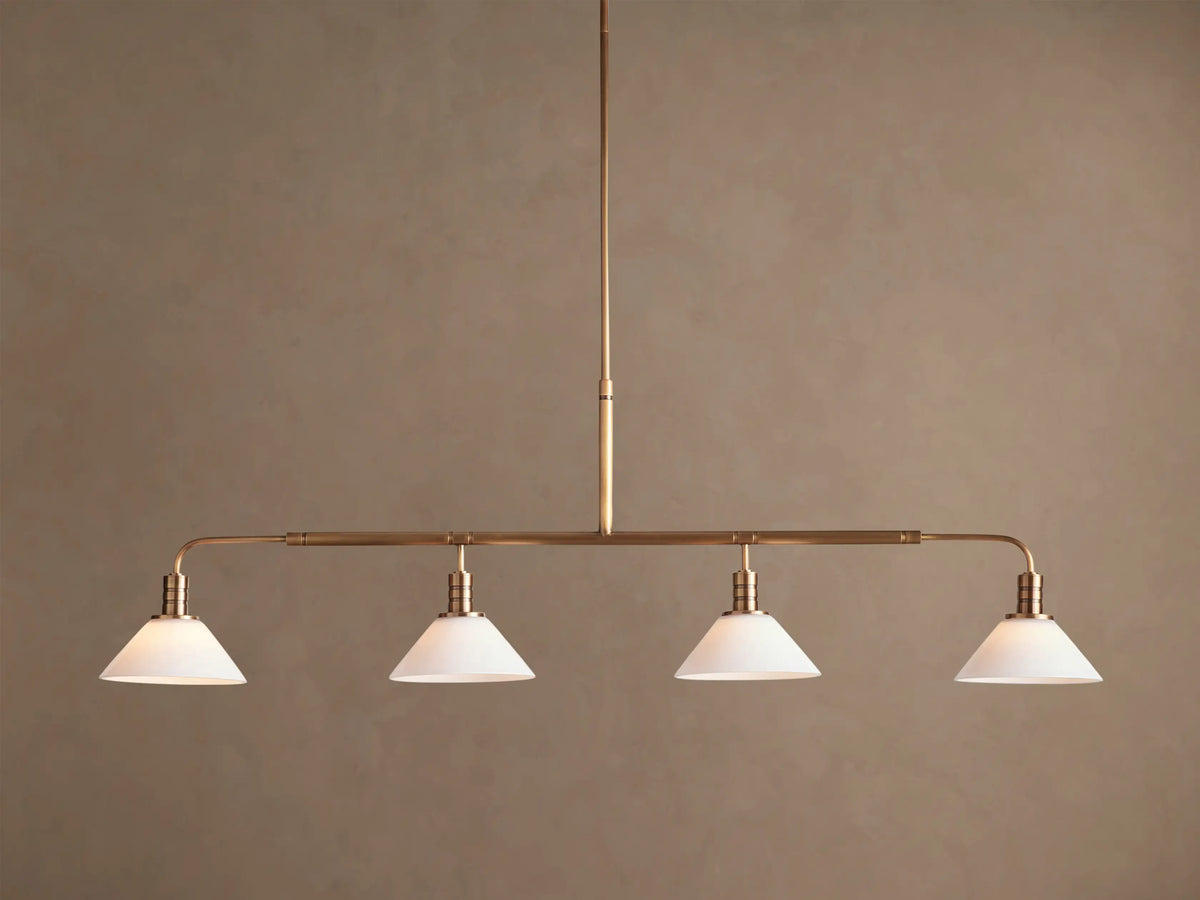 Theron Linear Chandelier with four milk glass cones on a horizontal brass bar, suspended against a warm brown wall.