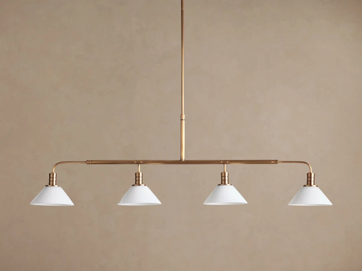 Theron Linear Chandelier with four milk glass cones on a horizontal brass bar, suspended against a warm brown wall.