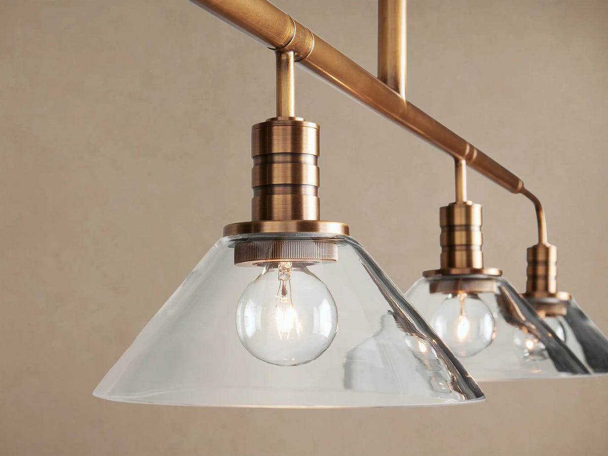 Theron Linear Chandelier, brass-toned with five frosted glass conical shades, hanging against a light brown wall.