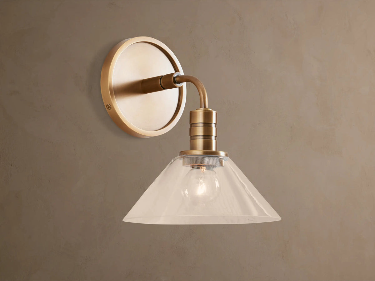 Theron Sconce, a brass wall-mounted light with a round backplate, curved arm, and clear glass cone shade, lit, on a taupe wall.