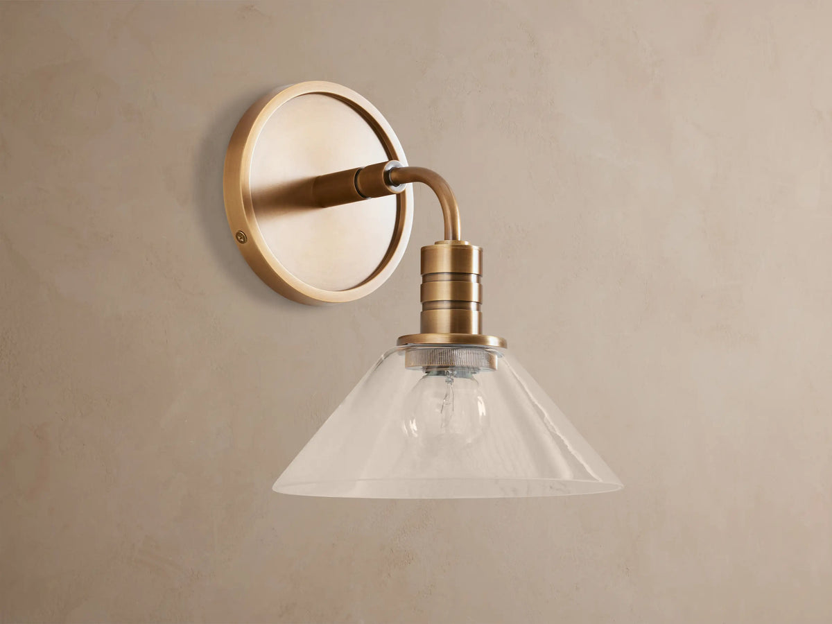 Theron Sconce, a brass wall-mounted light with a round backplate, curved arm, and clear glass cone shade, lit, on a taupe wall.