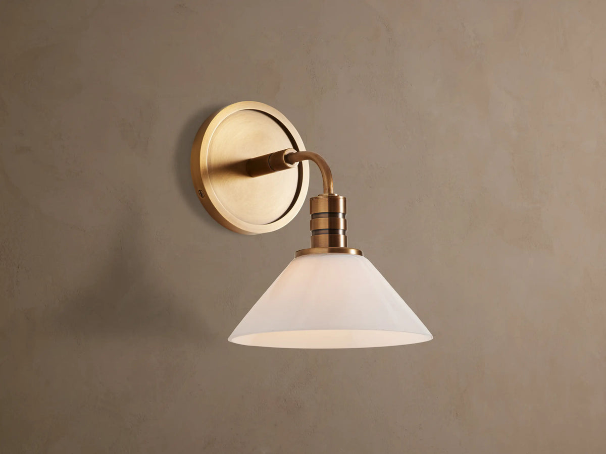 Theron Sconce with brass finish and white milk glass cone, mounted on a textured tan wall, emitting a soft glow.