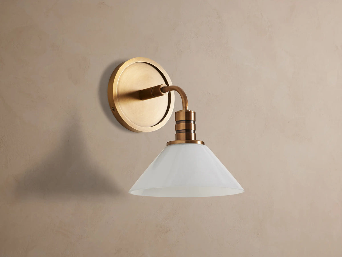 Theron Sconce with brass finish and white milk glass cone, mounted on a textured tan wall, emitting a soft glow.
