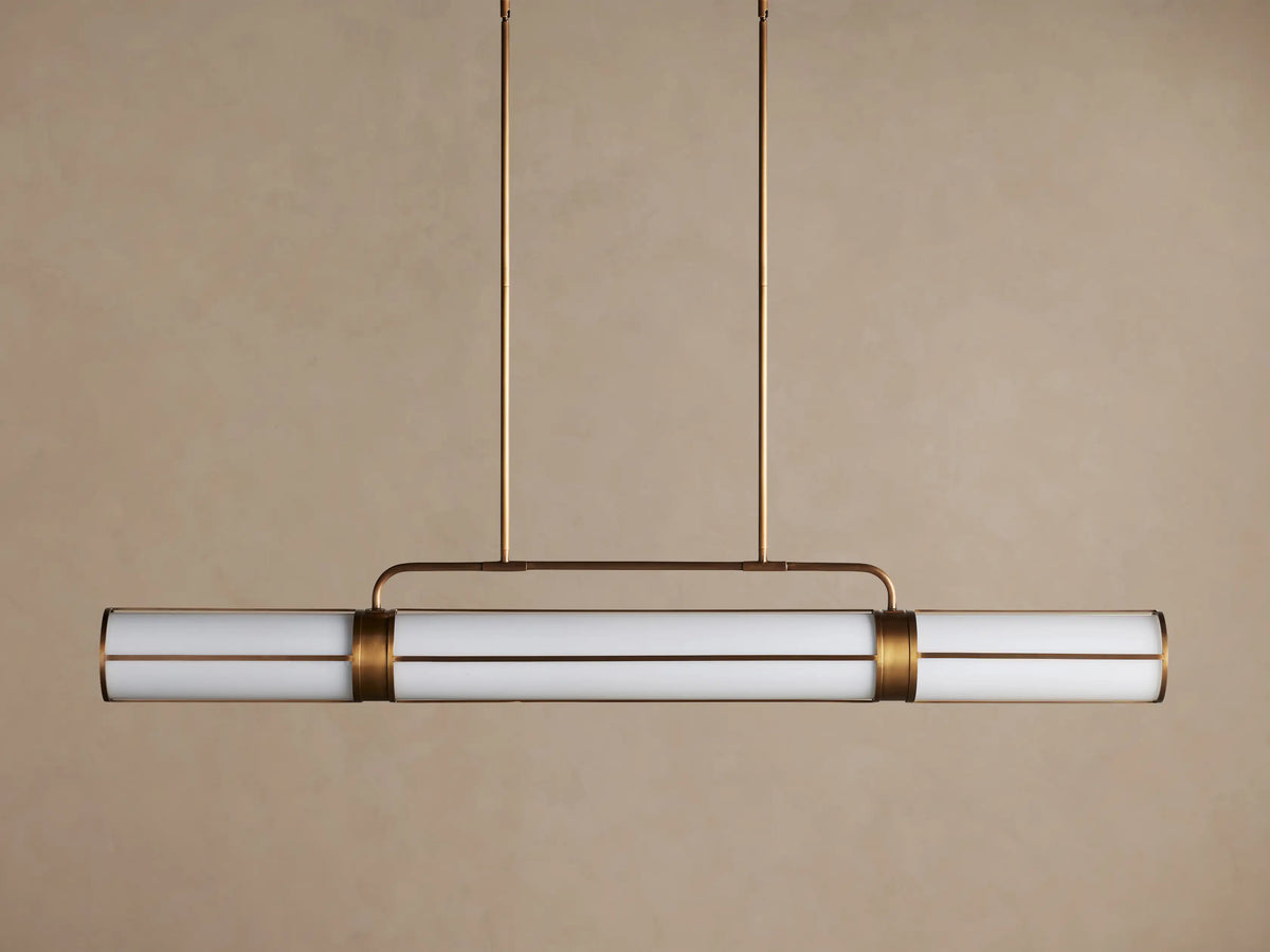 Remming Linear Chandelier with illuminated milk glass rods and burnished brass frame, hanging against a light brown wall.