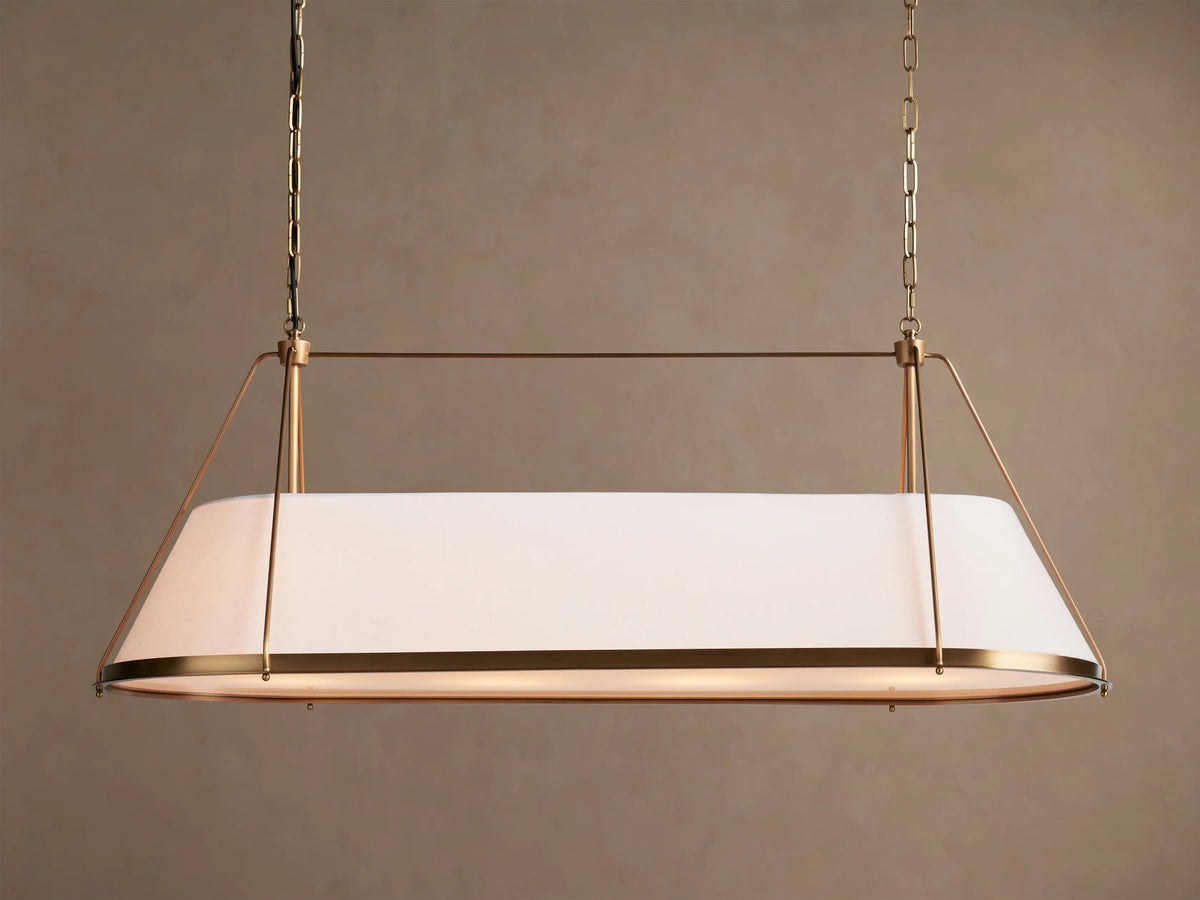 Shelton Linear Chandelier with burnished brass frame & illuminated tapered white linen shade, hanging from chains.