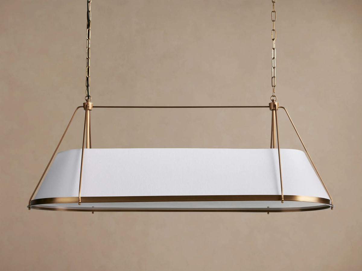 Shelton Linear Chandelier with burnished brass frame & illuminated tapered white linen shade, hanging from chains.