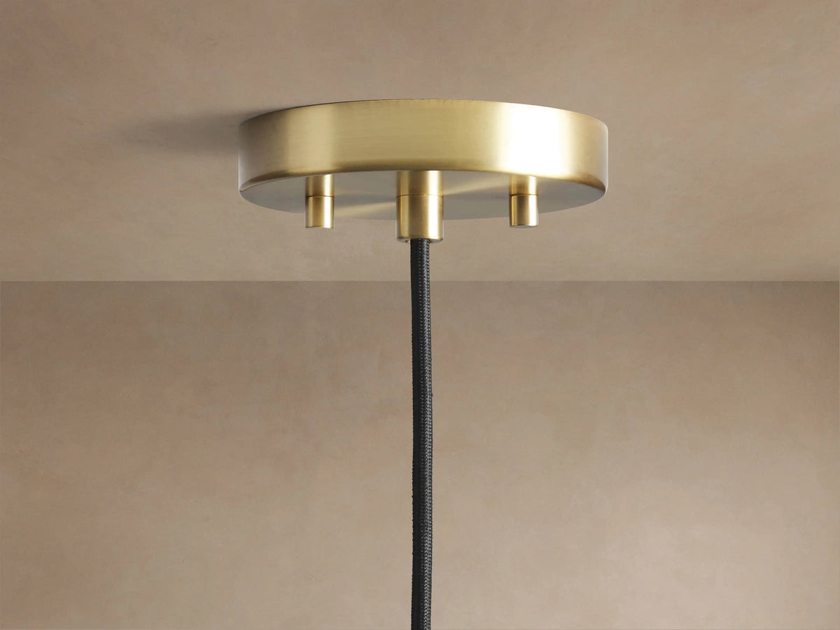 Organic 12" Pendant's round, brushed brass ceiling mount with a black fabric cord hanging from a light beige ceiling.