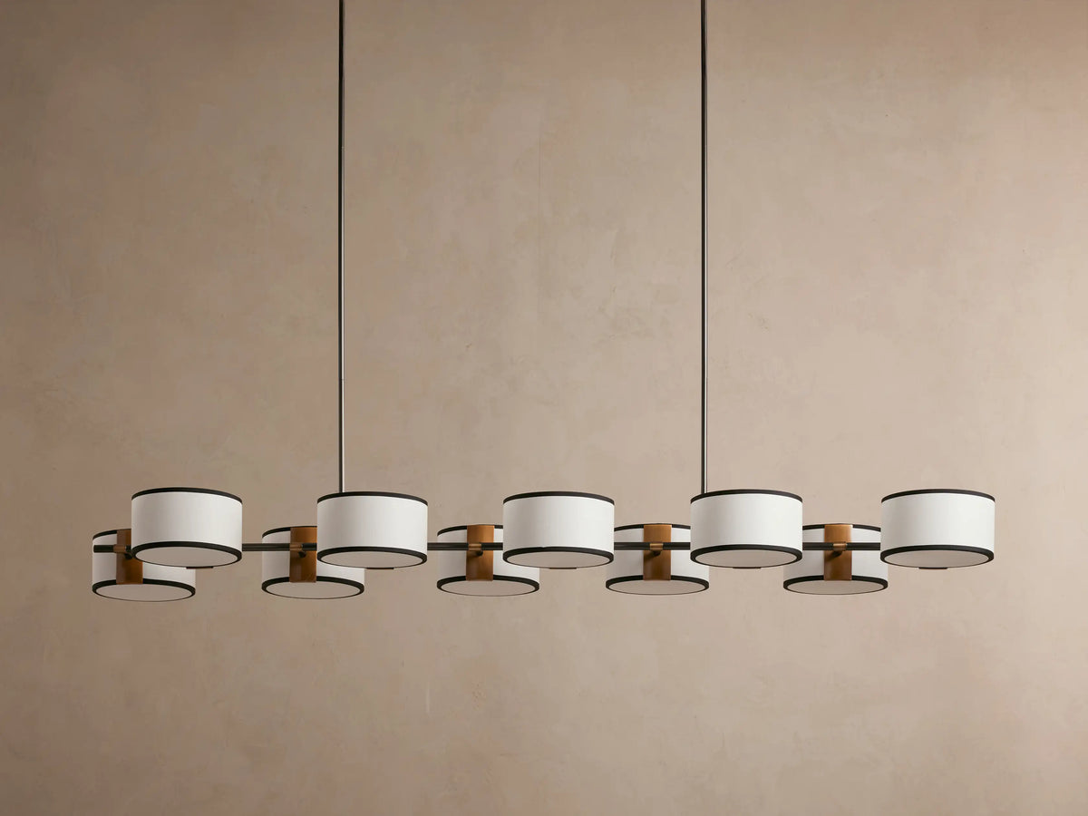 Hanging modern chandelier with ten cylindrical beige fabric lampshades on a dark metal frame against a light brown background.