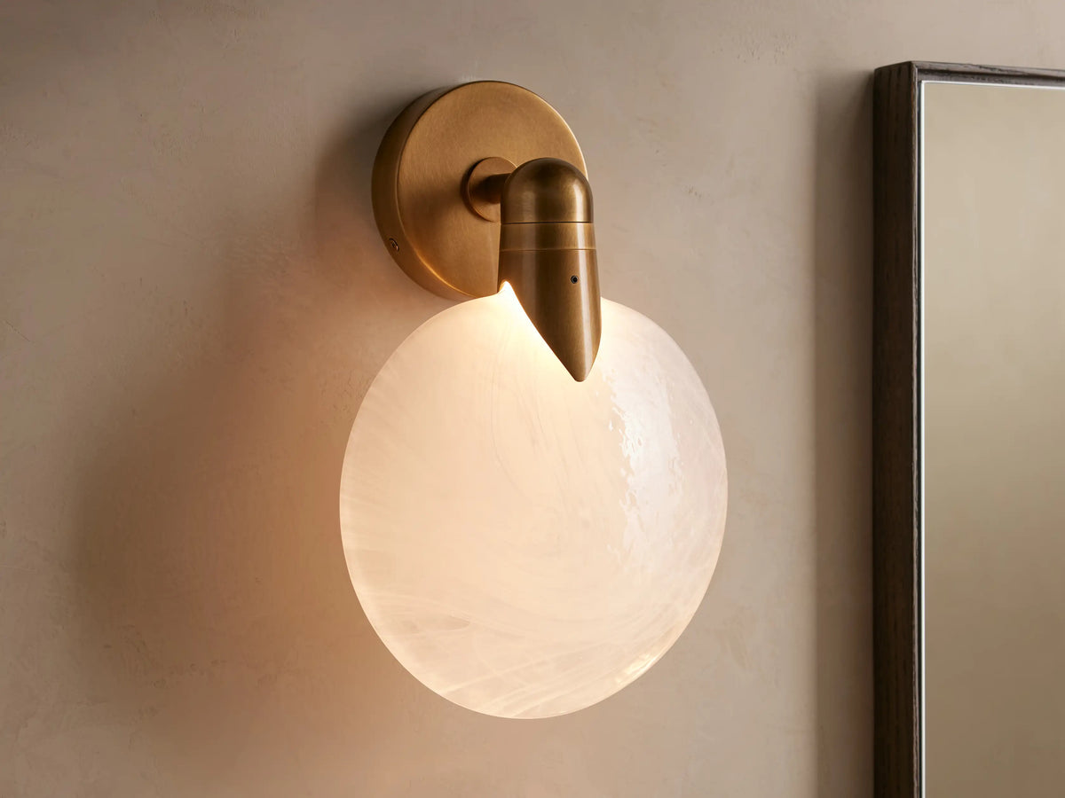 A hand-blown glass and brass Lyla Sconce with a round, frosted glass shade, mounted on a wall with a soft LED glow.