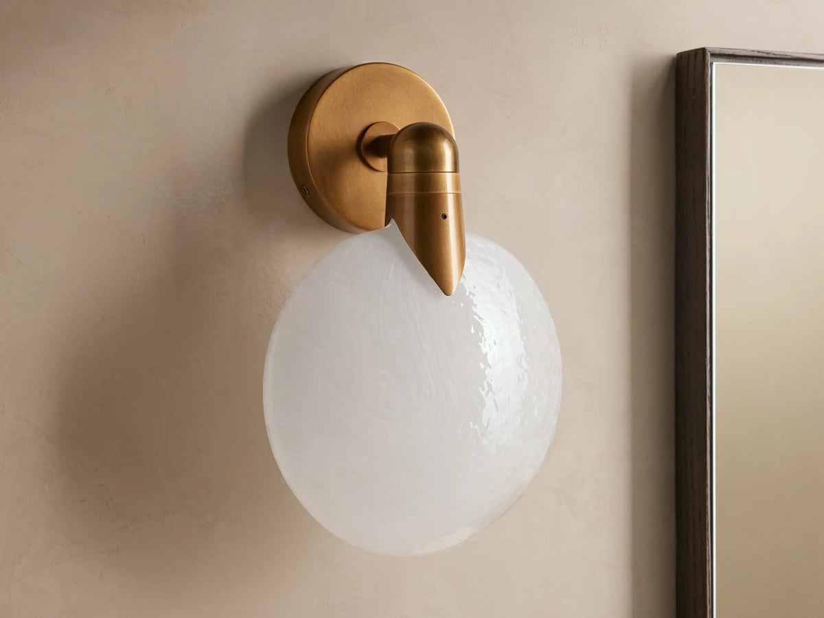A hand-blown glass and brass Lyla Sconce with a round, frosted glass shade, mounted on a wall with a soft LED glow.