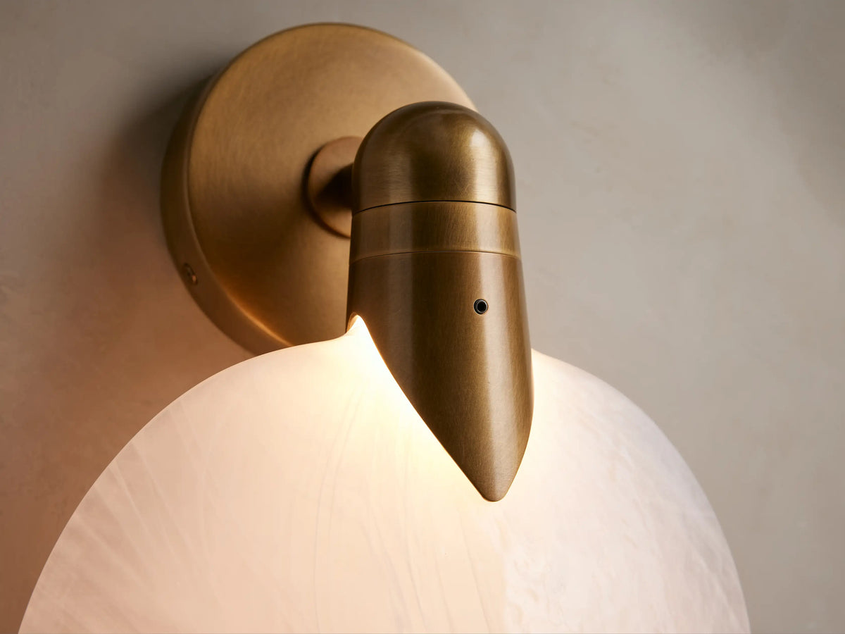 Lyla Sconce with a round, frosted glass shade and a brass-plated steel fixture mounted on a beige wall beside a framed mirror.