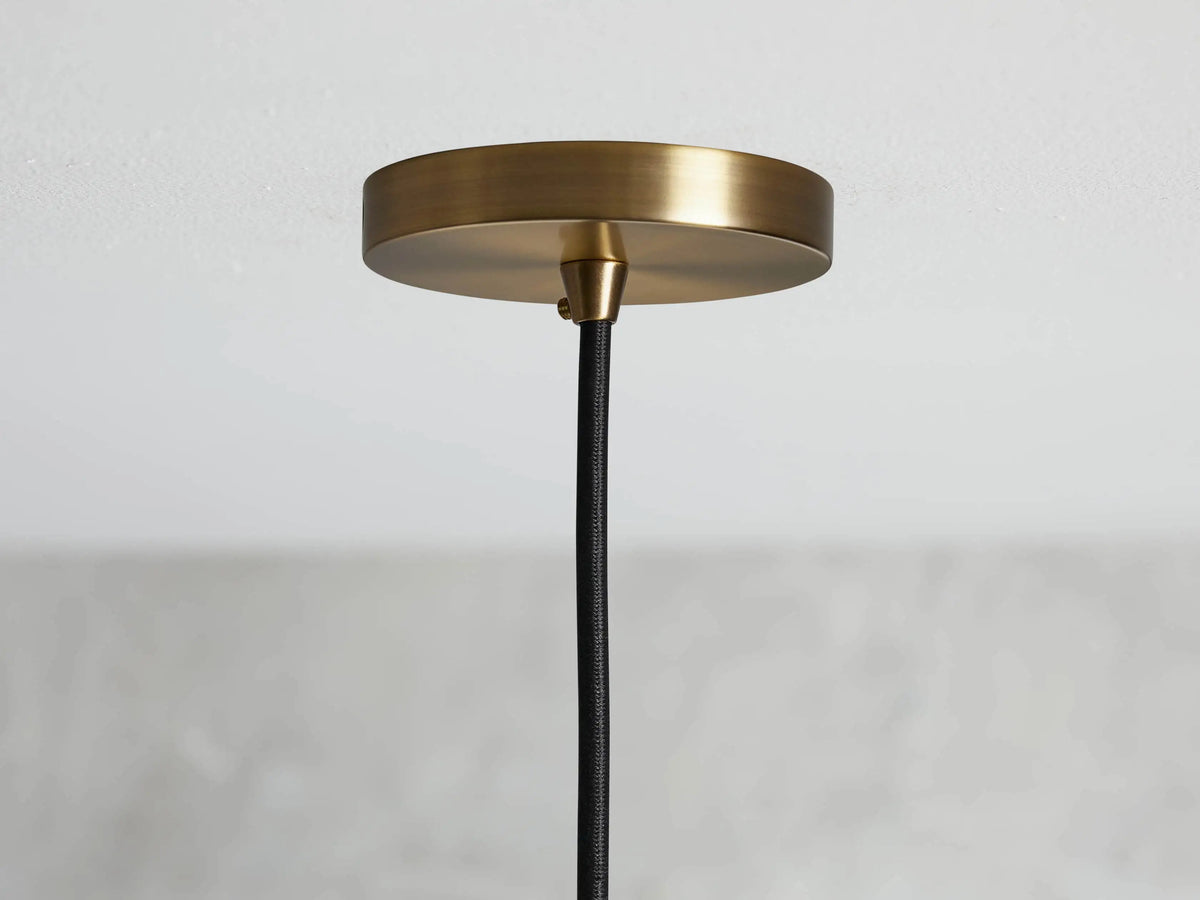 Ohlson Brass Swag Pendant featuring a brass-plated socket with a visible bulb, black fabric cord, and a gray blurred background.