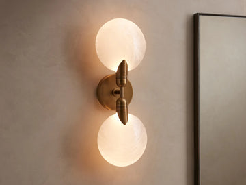 Lyla Double Sconce with two glowing spherical glass shades on a brass fixture, mounted vertically on a neutral wall.