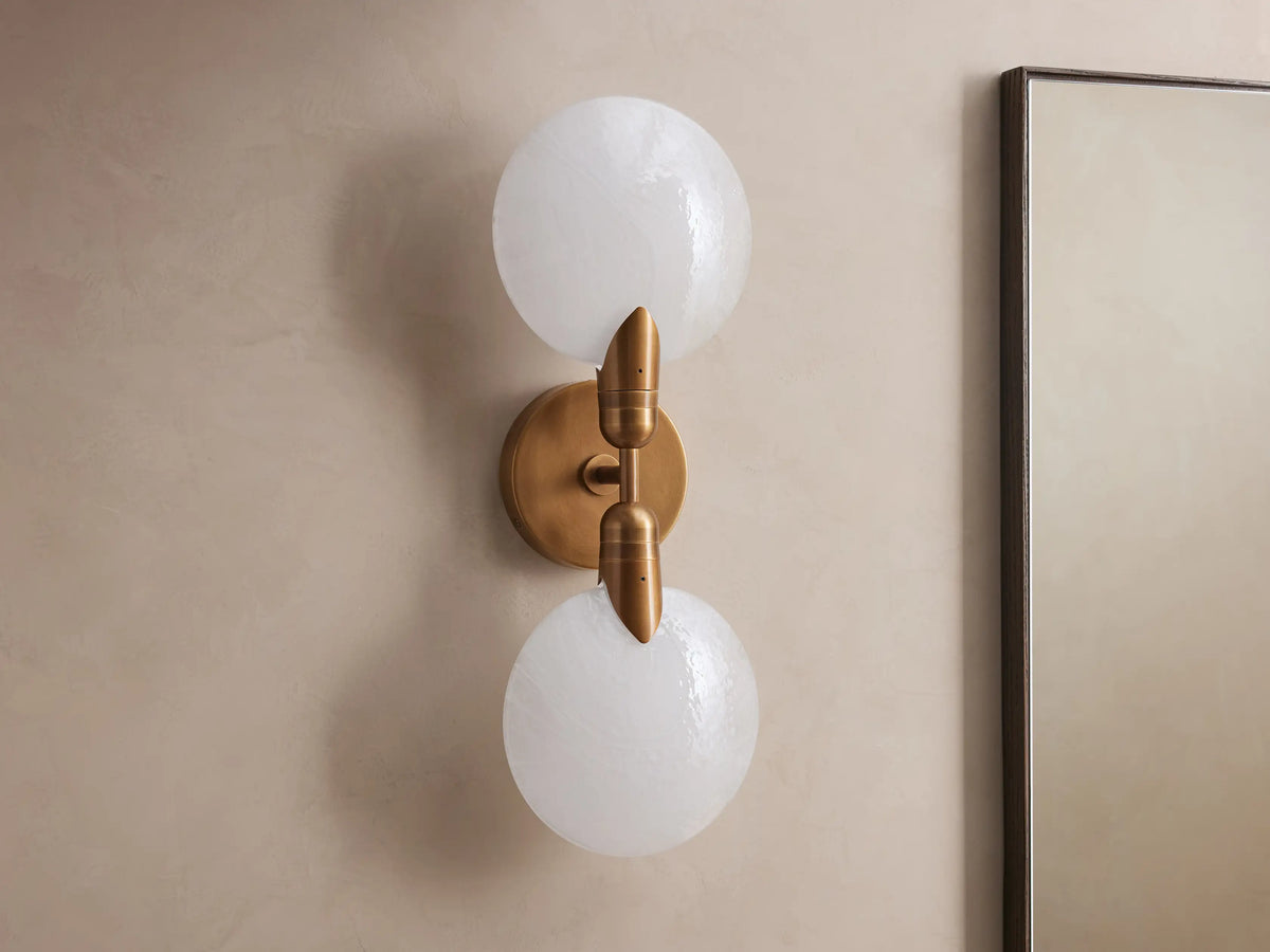 Lyla Double Sconce with two glowing spherical glass shades on a brass fixture, mounted vertically on a neutral wall.