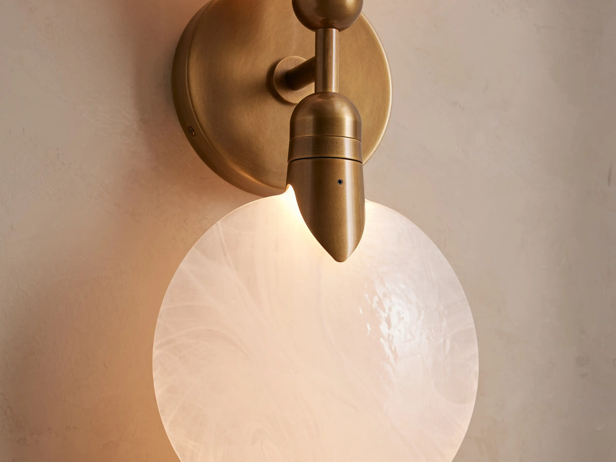 Lyla Double Sconce with two glowing hand-blown glass spheres mounted on a central brass steel backplate against a neutral wall.