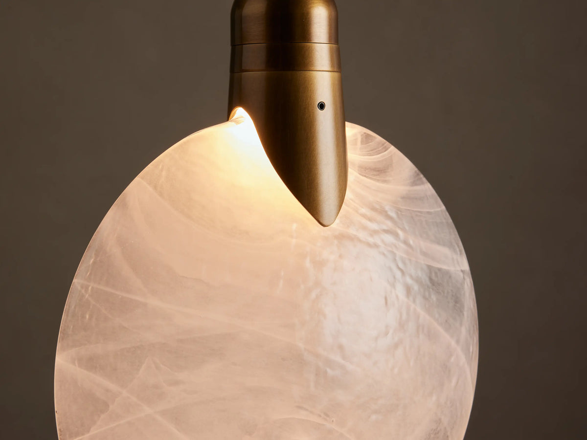 Lyla Single Pendant. A round, marbled white hand-blown glass shade hangs from a brass rod against a beige wall.