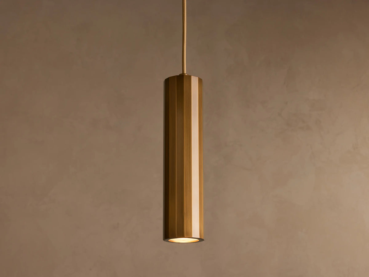 A cylindrical pendant light with a glossy brass finish hangs from the ceiling against a plain beige background.