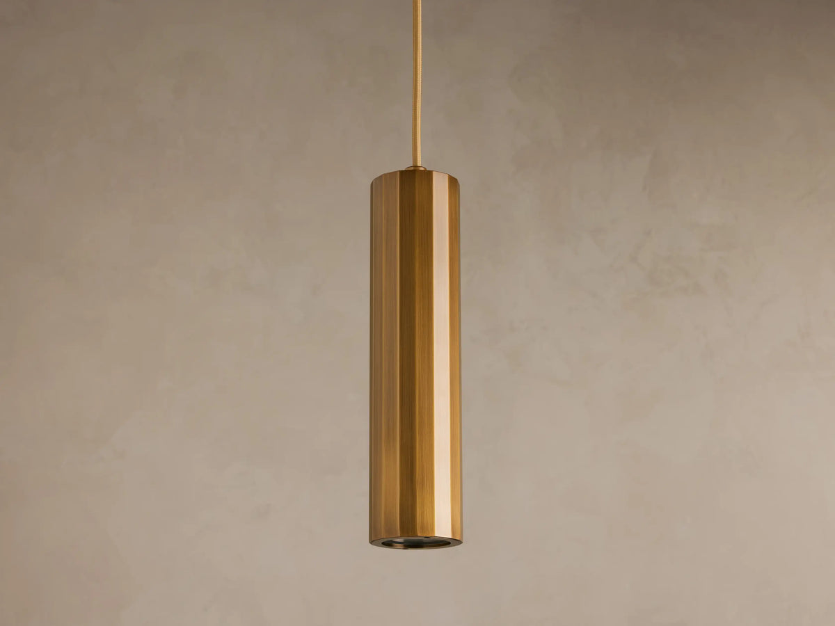 A cylindrical pendant light with a glossy brass finish hangs from the ceiling against a plain beige background.